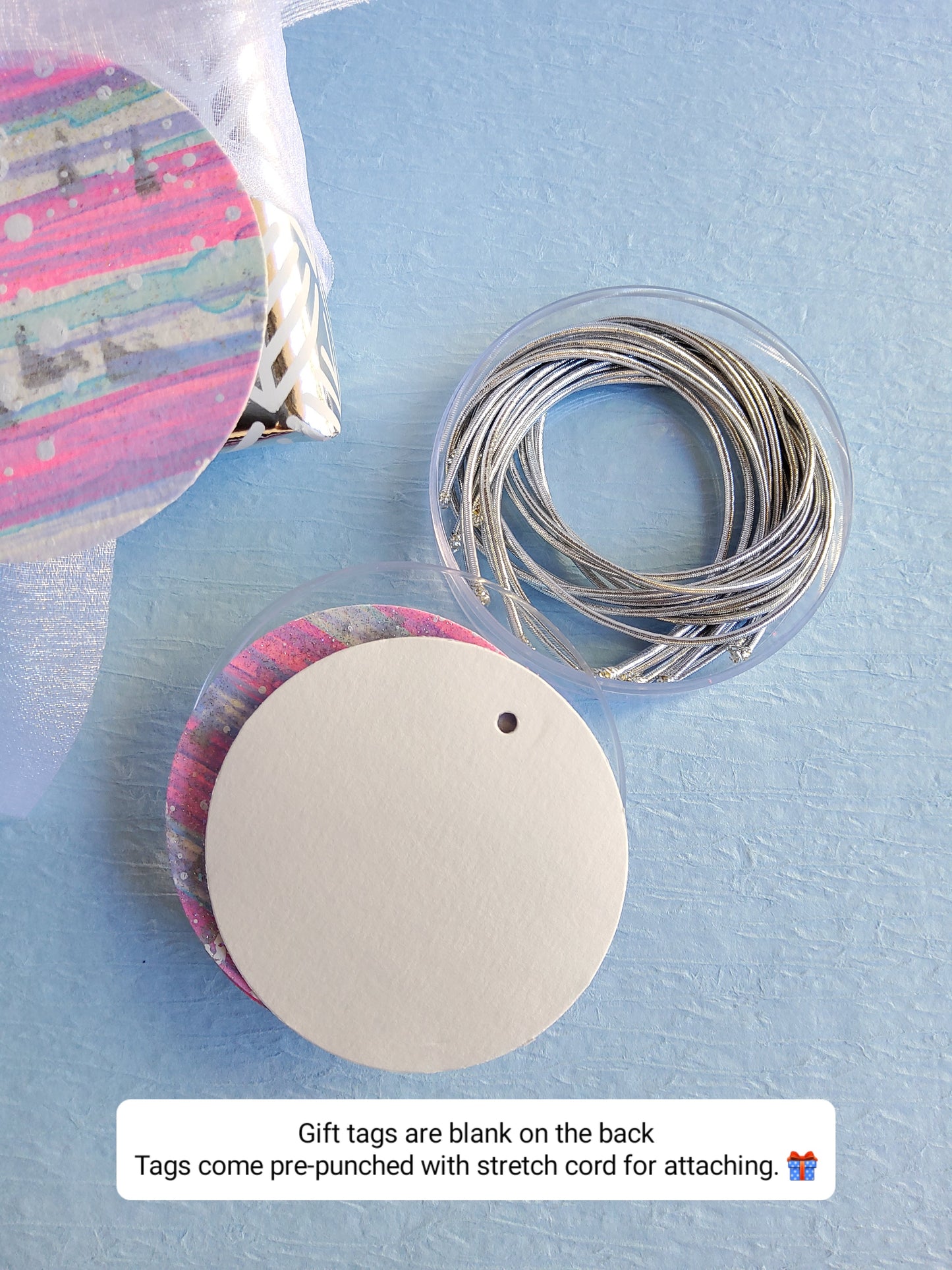 Set of hand-painted abstract gift tags with silver stretch cords displayed beside wrapped gift on light blue background