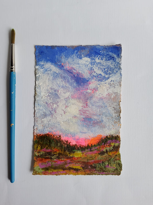 Vertical acrylic landscape with clouds, sunset tones, and deckle-edged handmade paper with paintbrush on a white background.