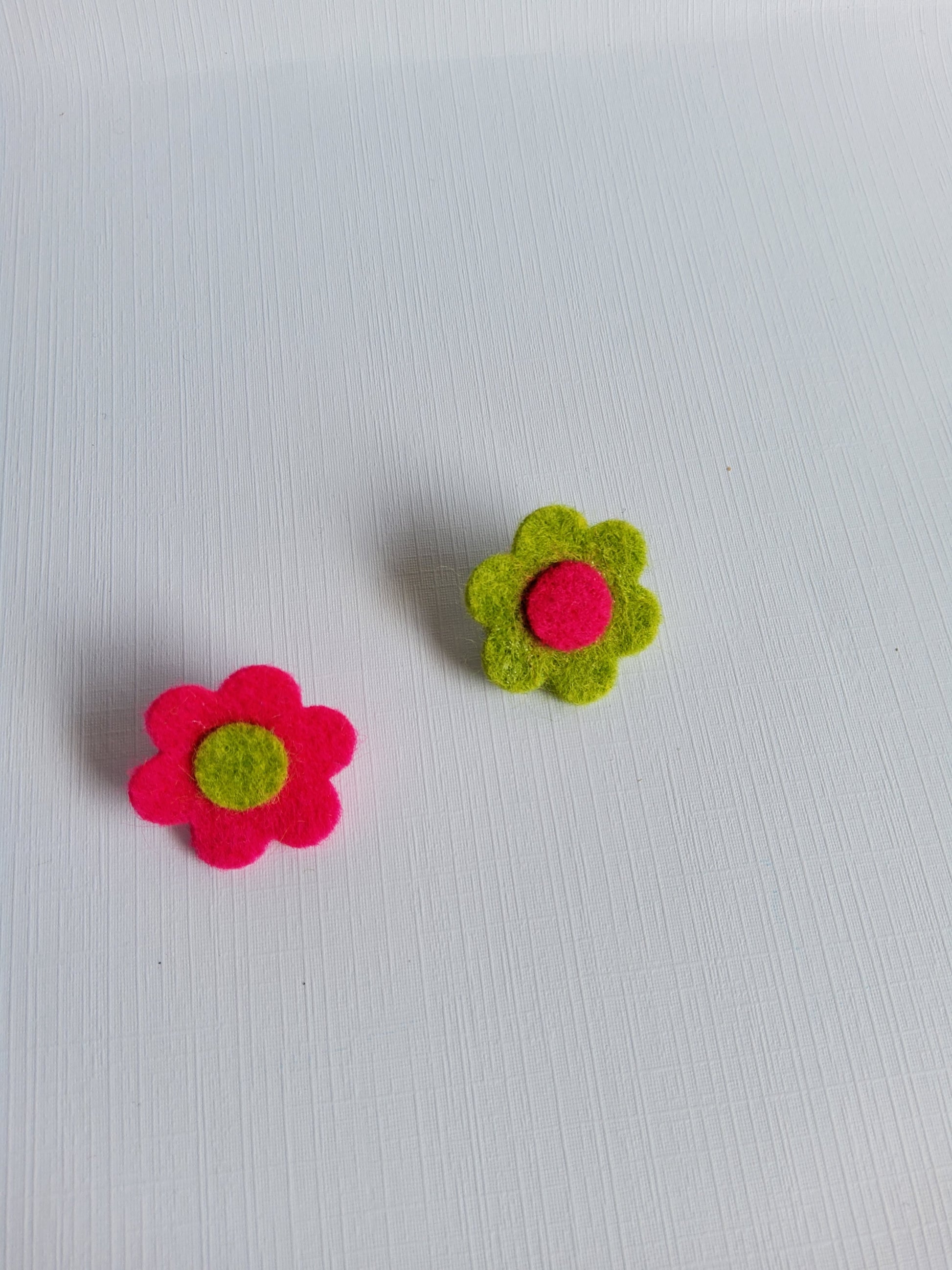 Two felt flowers, one green with a pink center and one pink with a green center, on a white background.