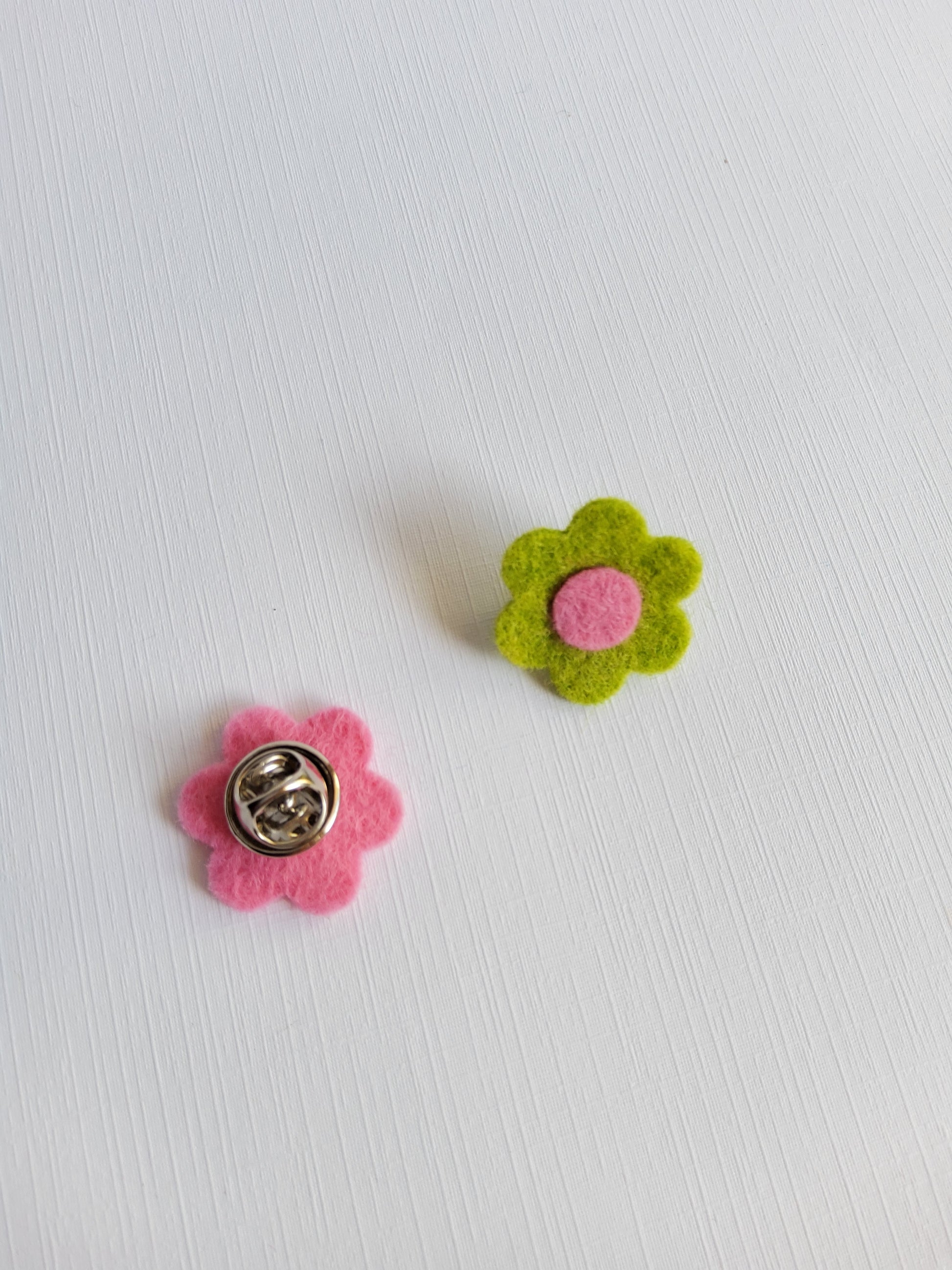 Two flower-shaped pins, one green with a pink center and the other pink with a silver clasp, on a textured white background.
