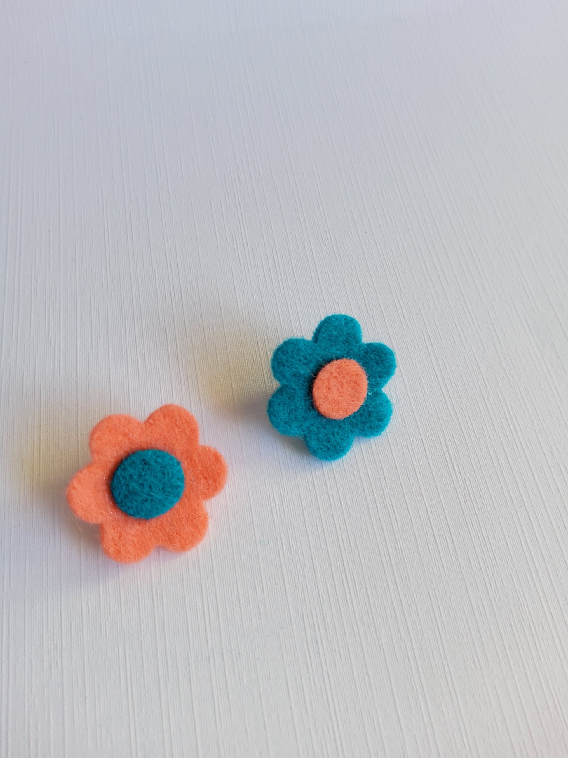 Two felt flowers, one blue with a orange center and one orange with a blue center, on a white background.