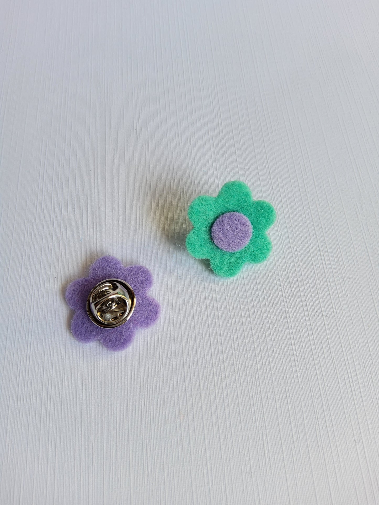 Two flower-shaped pins, one green with a purple center and the other purple with a silver clutch pin backing,  on a white background.