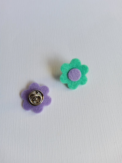 Two flower-shaped pins, one green with a purple center and the other purple with a silver clutch pin backing,  on a white background.