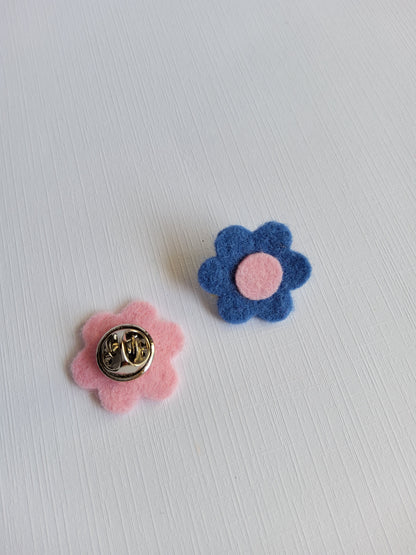 Two felt flower pins, one blue with a pink center and the other pink on a light gray background.