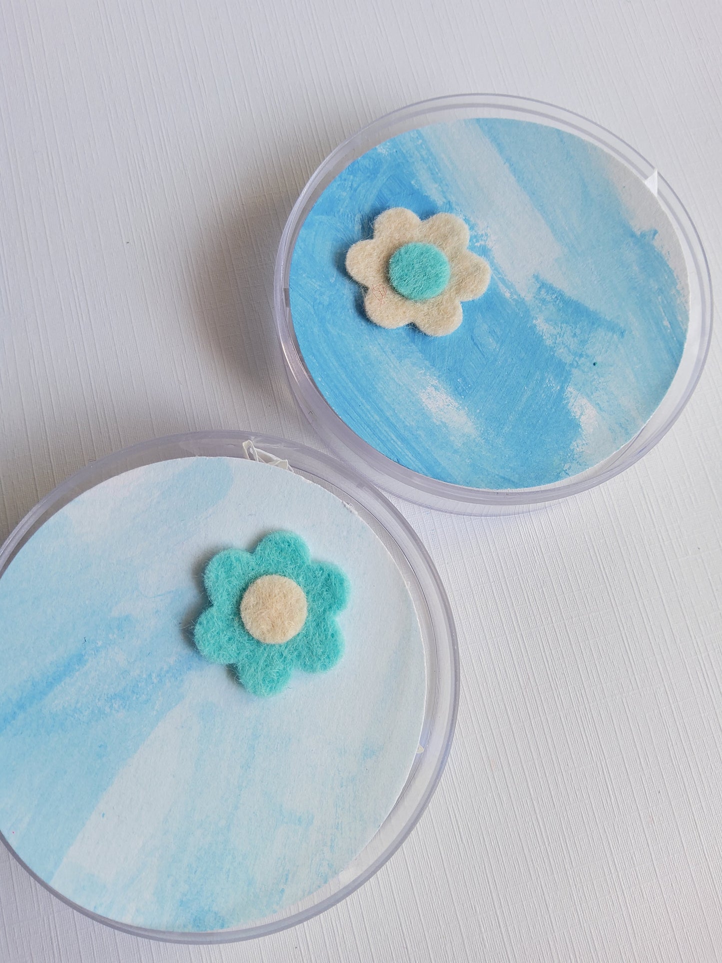 Two round containers with blue watercolor designs and flower-shaped lapel pins on a white background