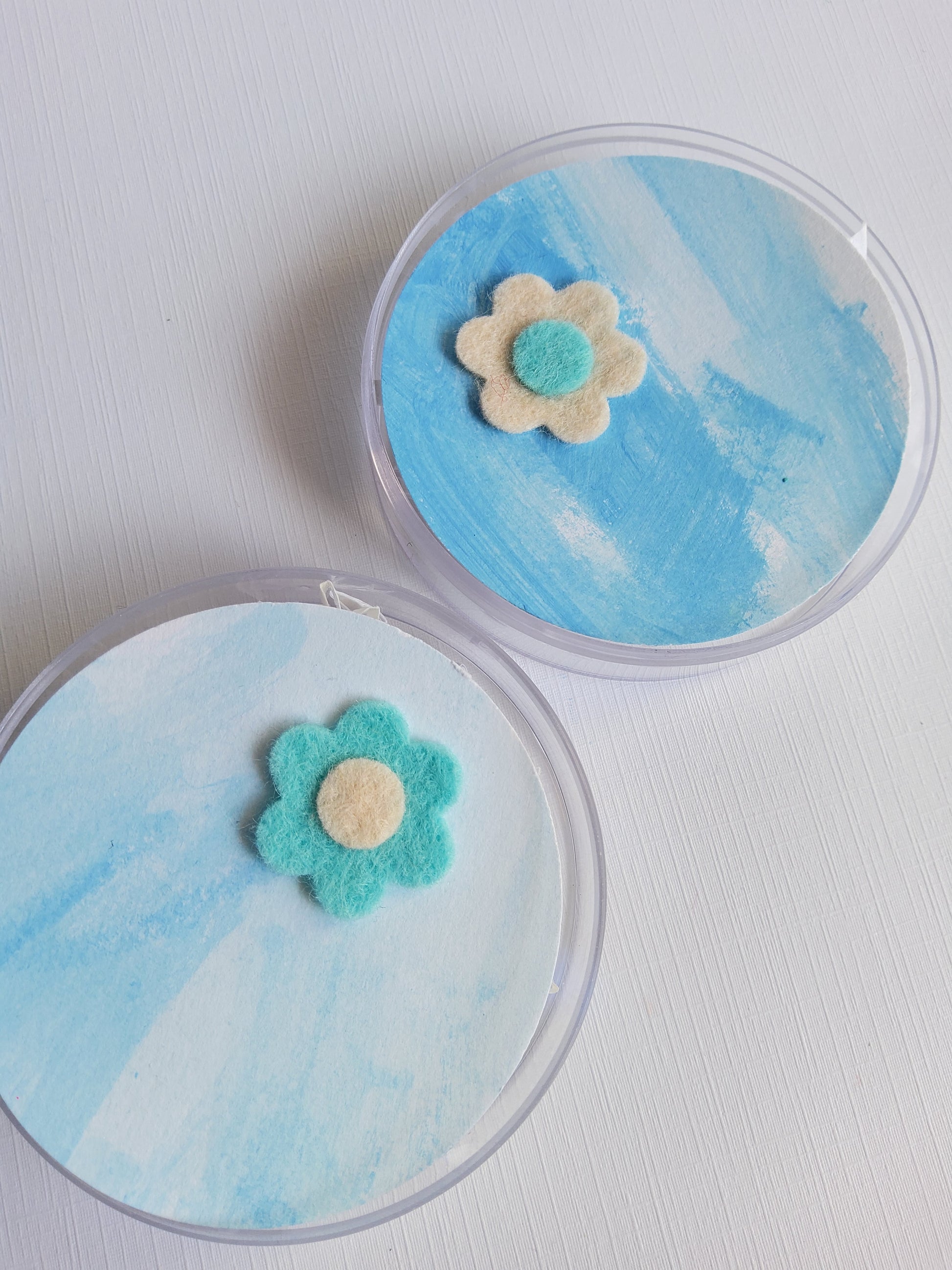 Two round containers with blue watercolor designs and flower-shaped lapel pins on a white background