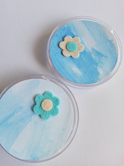 Two round containers with blue watercolor designs and flower-shaped lapel pins on a white background