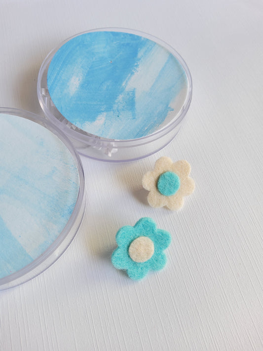Two round containers with painted blue tag and two decorative flower pins  on a light background