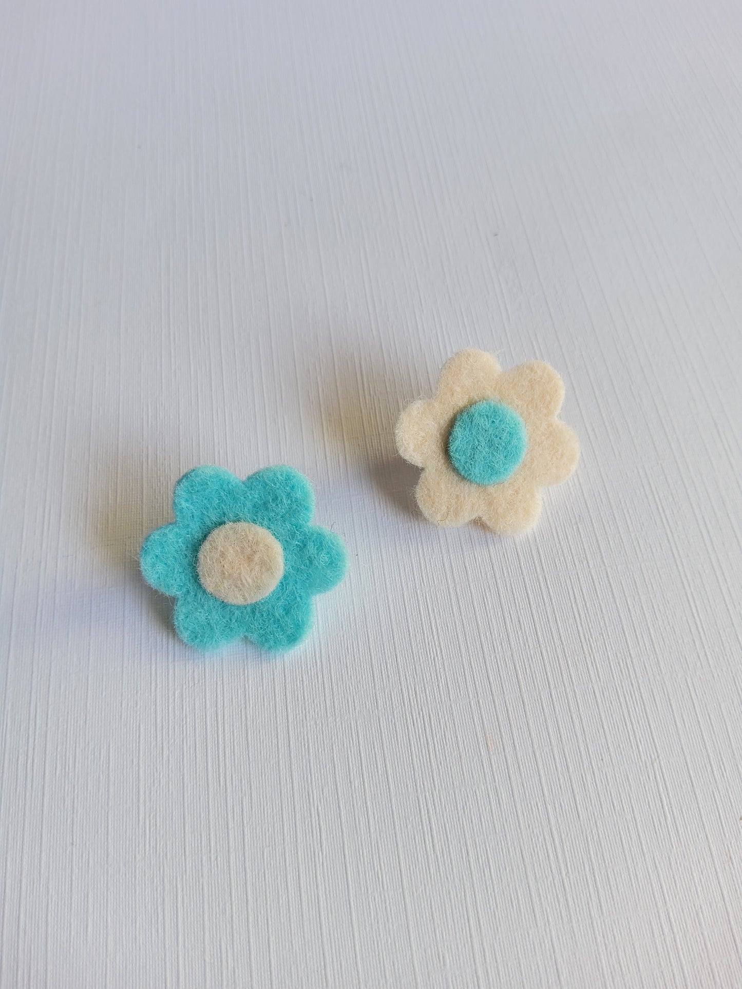 Two felt flower pins, one white with a blue center and the other blue with a white center, on a light gray background.