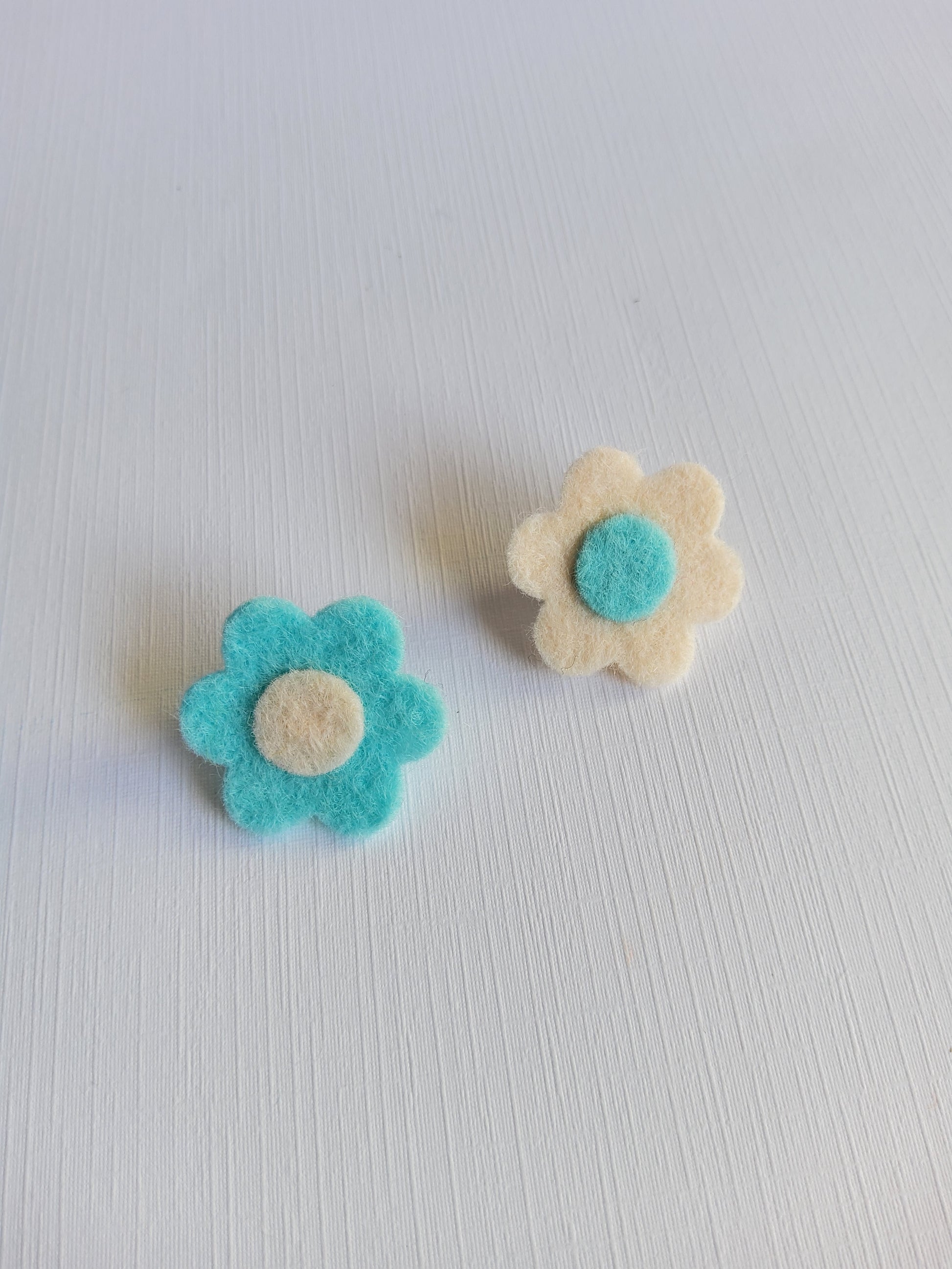 Two felt flower pins, one white with a blue center and the other blue with a white center, on a light gray background.