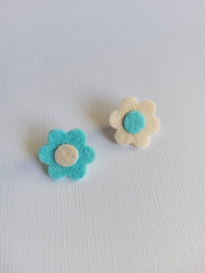 Two felt flower pins, one white with a blue center and the other blue with a white center, on a light gray background.