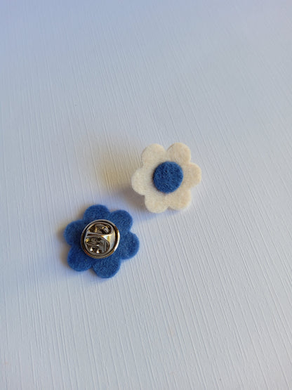 Two flower-shaped pins, one white with a blue center and the other blue showing the back clutch closure , on a light gray background.