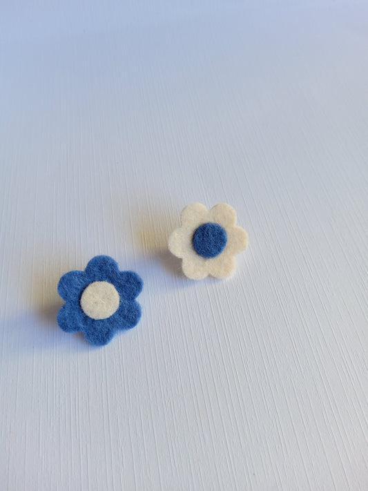 Two felt flower pins, one white with a blue center and the other blue with a white center, on a textured light gray background.