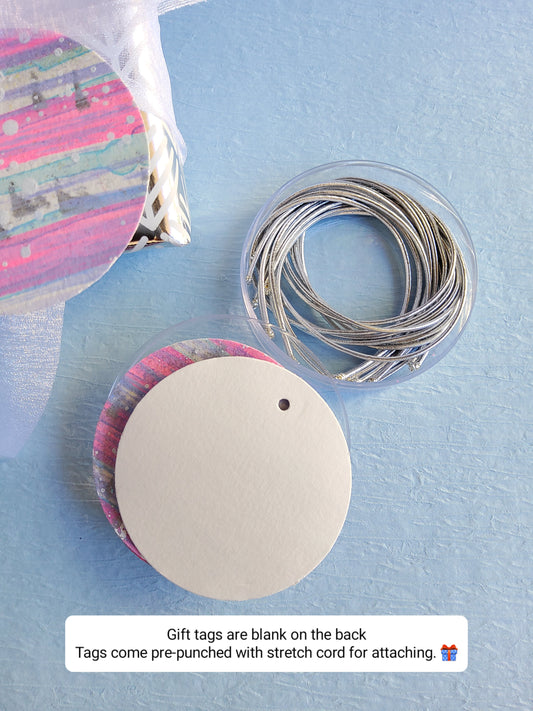 Set of hand-painted abstract gift tags with silver stretch cords displayed beside wrapped gift on light blue background 