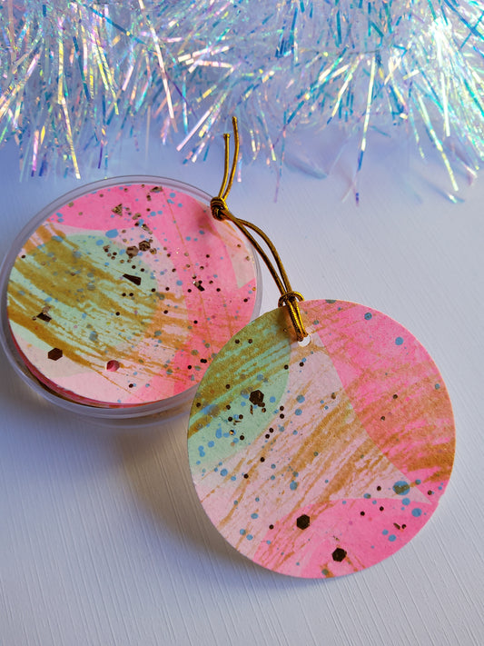 Pink and green hand-painted abstract gift tags with silver stretch cords displayed beside iridescent tinsel on white background