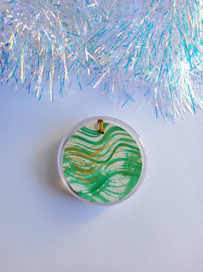 Green  and gold abstract hand-painted round gift tags with gold cords in a clear round acrylic container beside iridescent tinsel background