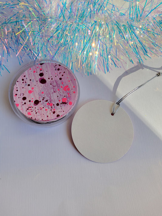 Purple & neon pink abstract hand-painted round gift tags with silver cords in a clear round acrylic container beside iridescent tinsel on a white background