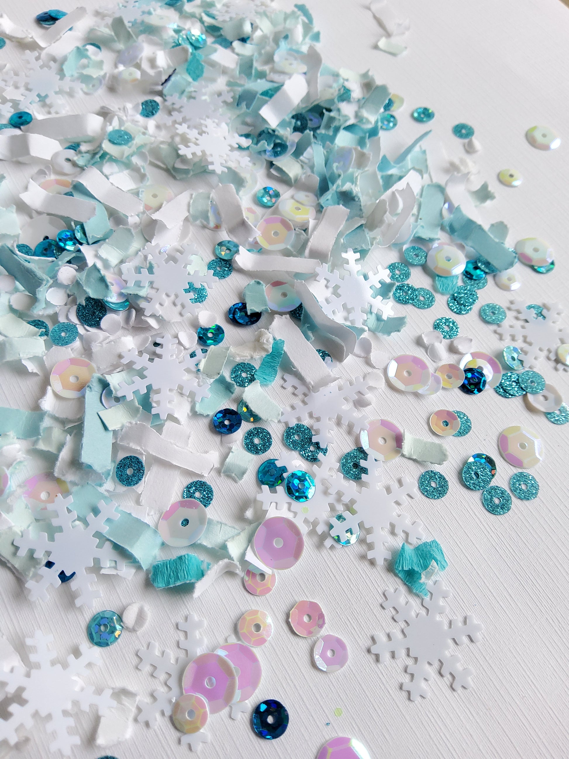 Turquoise and white paper confetti with iridescent white sequins,  snowflake shape embellishments, and turquoise sparkly sequins on white background. 