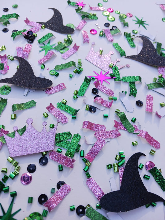 Pink glitter crown, black glitter witch hat with pink and green glitter confetti and sequins on a white background. 